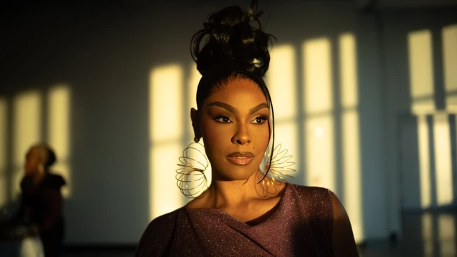 Woman with hair in a high bun and sculptural wire hoop earrings, lit by warm sunlight streaming through windows onto a wall behind her.