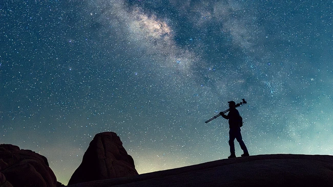 Silhouette of a person carrying a tripod over one shoulder, standing on a rock ridge beneath a bright Milky Way sky.