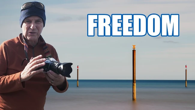 Photographer holding a camera on a beach with wooden pilings and calm water, with text overlay reading 'FREEDOM'