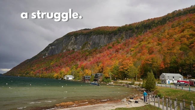 Fall foliage in red and orange covers a steep cliff rising above a lake, with lakeside cabins and a person standing on the shore.