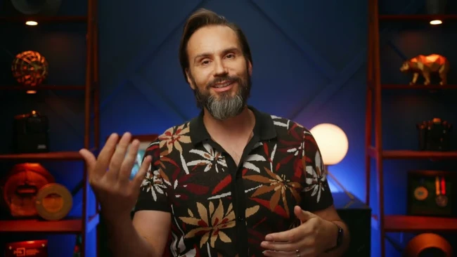 Smiling bearded man in a floral short-sleeve shirt gesturing with both hands in front of a blue-lit shelf set.