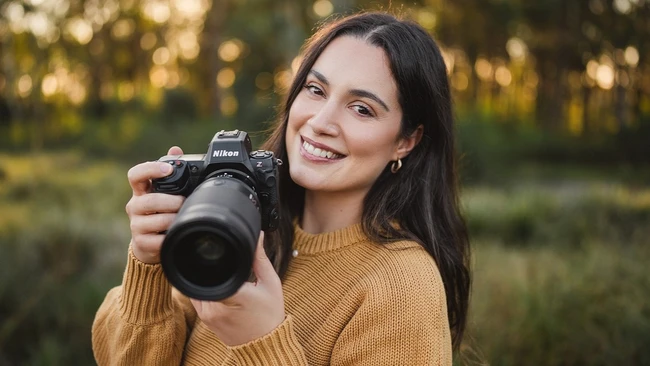 Smiling woman with long dark hair in a mustard sweater holding a Nikon mirrorless camera with a large zoom lens, outdoors in soft light.