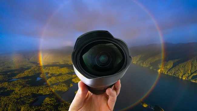 Hand holding a wide-angle lens with a rainbow halo around it, in front of an aerial view of forested hills and a lake