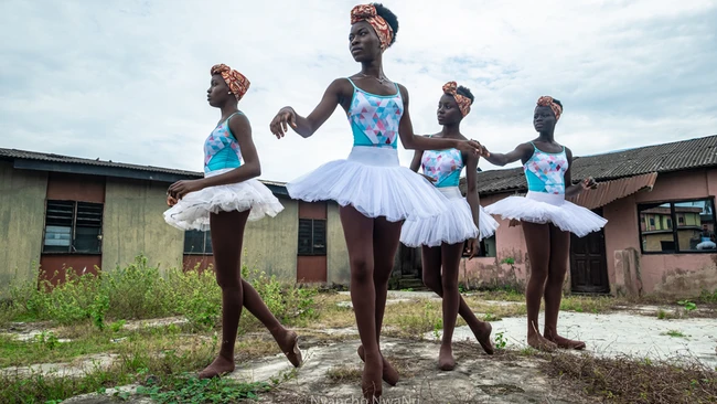 Four young dancers in white tutus and floral leotards with orange head wraps posing outdoors before weathered buildings.