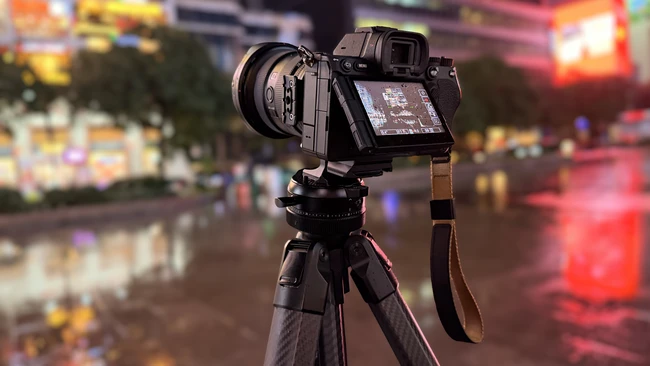 Mirrorless camera with a lens mounted on a carbon fiber tripod, rear screen angled out, set on a wet city street at night.