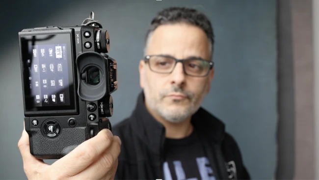 Man in glasses and a black jacket holding up a black mirrorless camera toward the viewer, its rear screen showing menu icons.