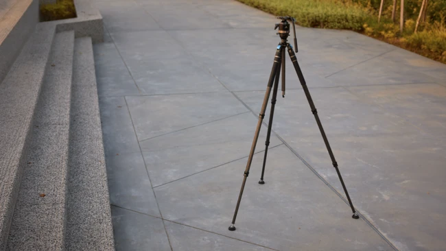 Dark tripod with a fluid video head and panning handle standing on a concrete plaza beside granite steps