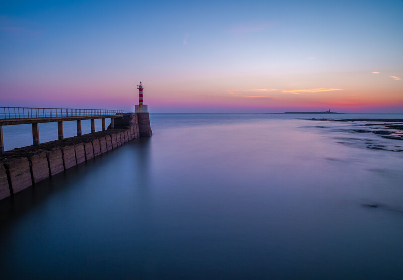 Lighthouse at the end of a long pier extending into calm water at dusk, with purple and blue twilight sky.
