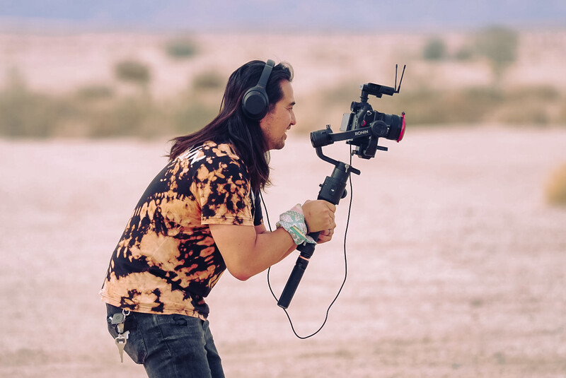 Videographer operating a gimbal-stabilized camera rig while filming outdoors near water.