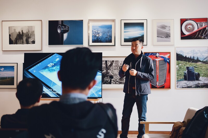 a man giving a lecture to a classroom, with photographs hanging behind him