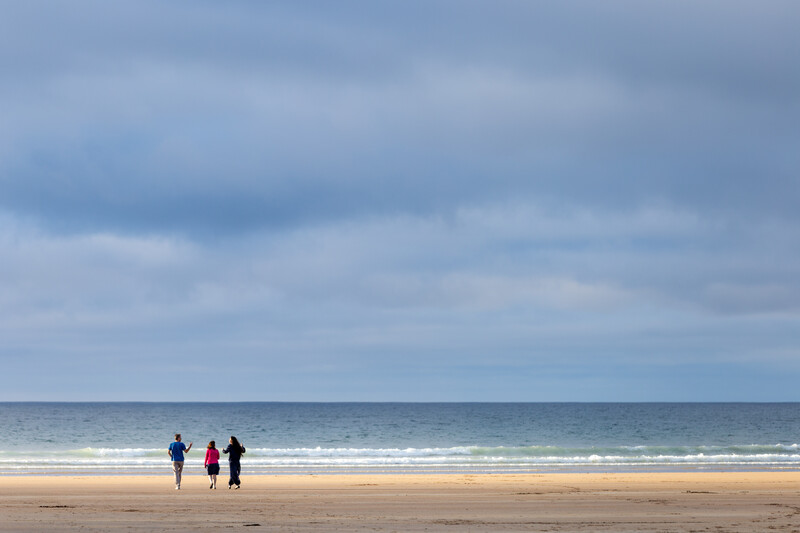 Three people walk on a sandy beach towards the horizon above the sea.