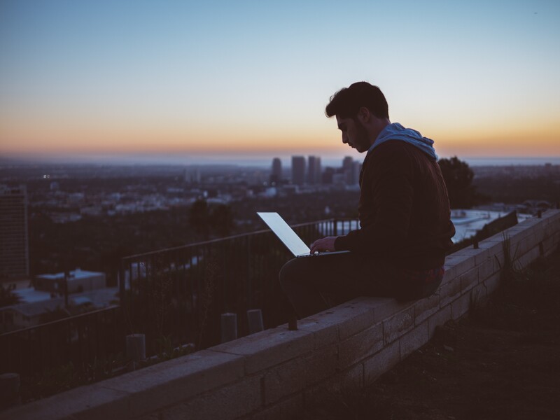 a man on his laptop, sitting on a ledge, overlooking a city 