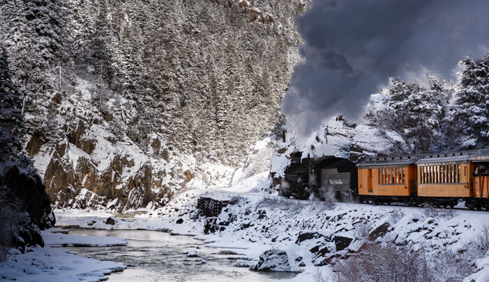 Photograph of a steam train traveling along mountain river in the snow.