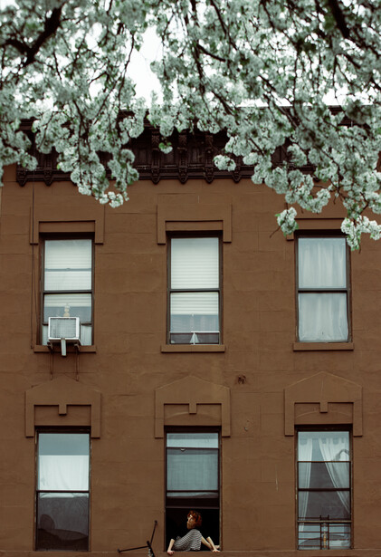 A young woman sits on her Brooklyn window sill for the Come To My Window project
