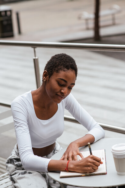 A young woman writing on a notebook at an outdoors cafe. 