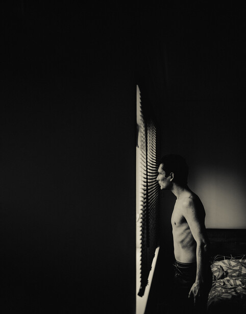 A topless man looking through the window blinds.