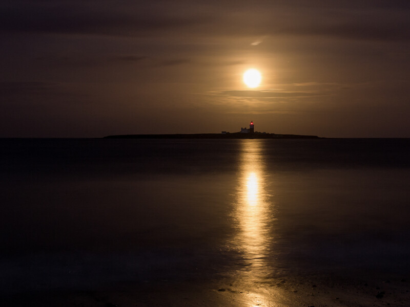 Full moon reflected in calm water with silhouetted ship on horizon.