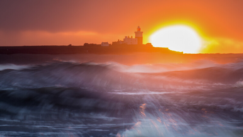 Coastal cityscape silhouetted against a brilliant sunset over turbulent ocean waves.