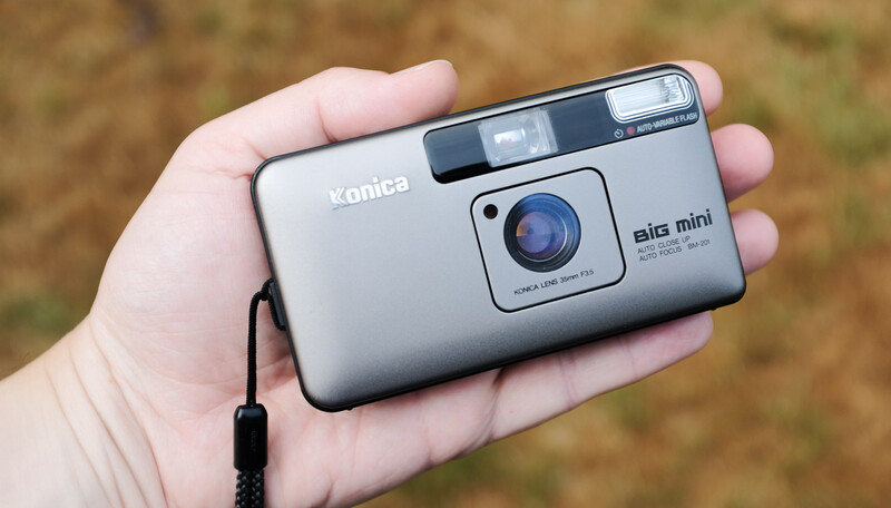 Konica Big mini compact film camera held in palm of hand, showing silver body, blue lens, and built-in flash.