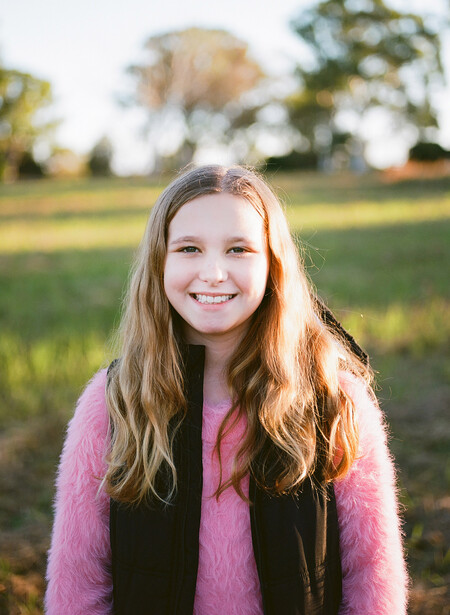 Young woman with long reddish-brown hair smiling at camera in an outdoor field during golden hour.