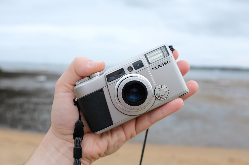 Hand holding a silver rangefinder camera with a blue lens element on a beach.