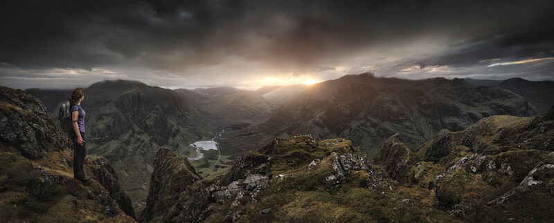 Hiker standing on rocky outcrop overlooking a mountain valley with a river and dramatic sunset sky.
