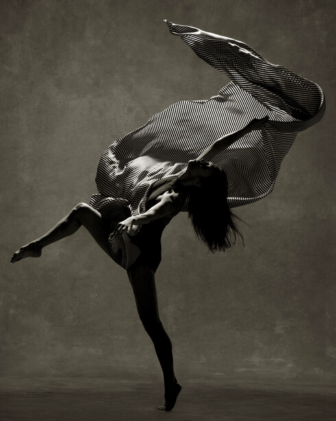 Female dancer captured mid-leap against neutral background, body inverted with legs extended upward.