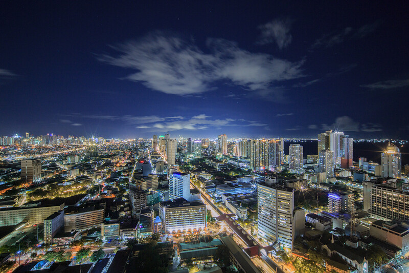 Aerial cityscape at dusk with illuminated high-rise buildings and streets stretching toward distant mountains.