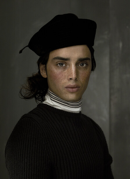 Portrait of a man wearing a black beret and ribbed sweater with white collar against neutral background.