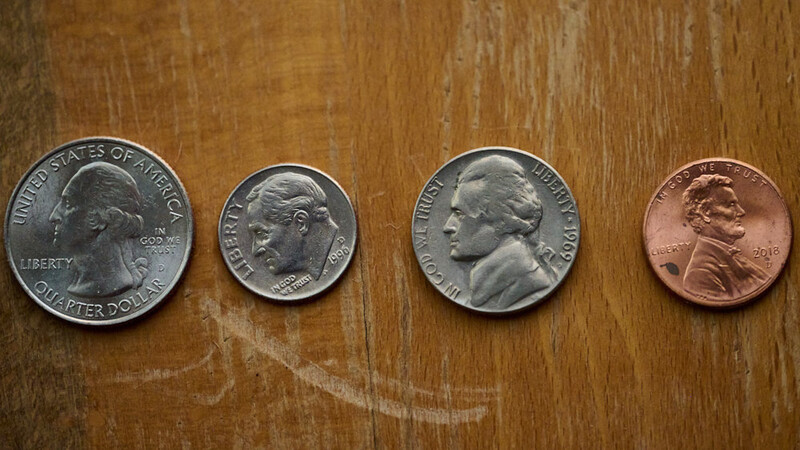 Four U.S. coins arranged in a row on a wooden surface: a quarter dollar, dime, quarter, and penny.