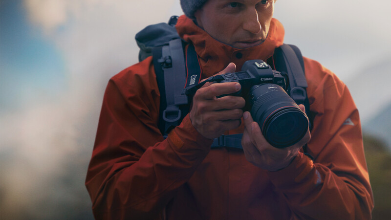 Photographer in orange jacket holding a telephoto lens camera against a mountainous backdrop.