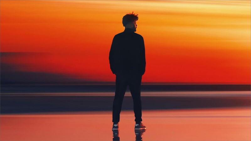 Silhouetted figure standing on a reflective surface against a vibrant orange and red sunset sky.