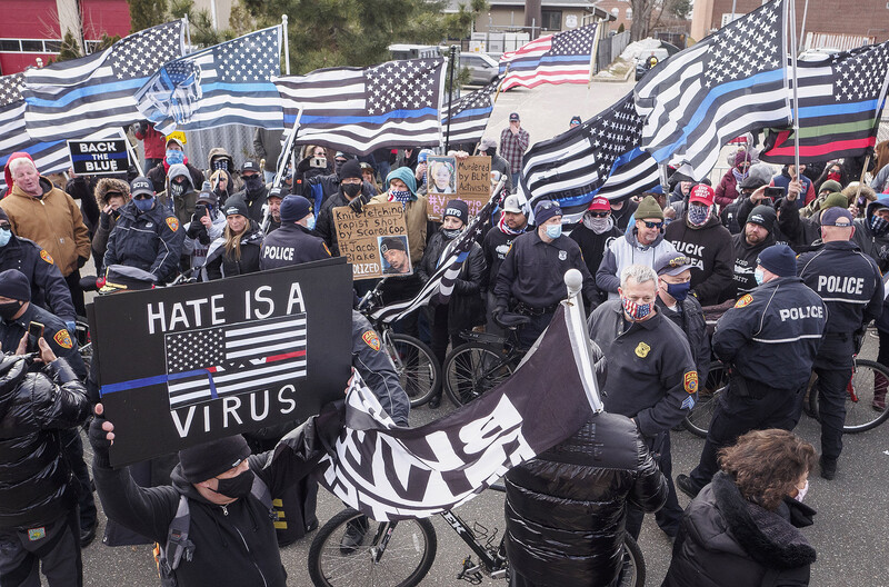 Large outdoor gathering with police presence, protest signs, and flags with thin blue line imagery.