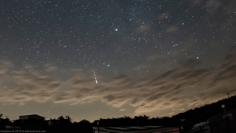 Night sky filled with stars above a silhouetted landscape with clouds and a bright meteor streak.
