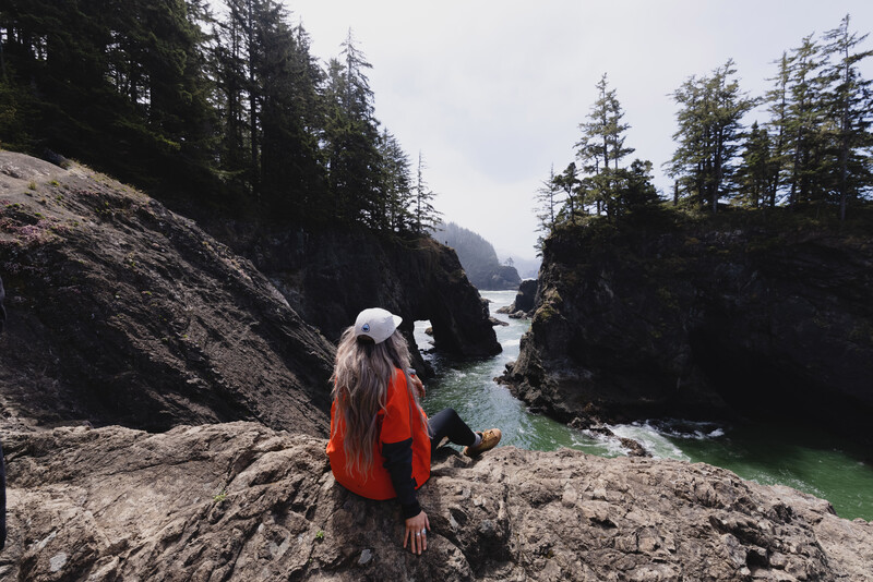 Hiker in red jacket perched on rocky outcrop overlooking turquoise water and forested coastline.
