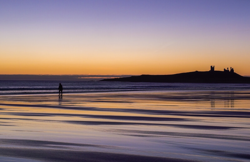 Solitary surfer standing in shallow water at sunrise, facing calm ocean waves with silhouetted headland in background.