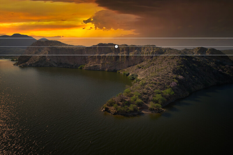Aerial view of a forested peninsula surrounded by calm water at sunset, with dramatic golden and orange sky.