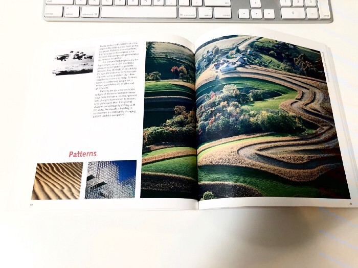 Open photography book displaying aerial landscape images alongside a keyboard and reference materials on a white desk.