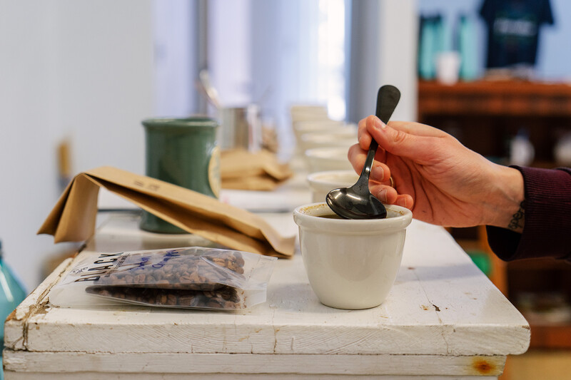 Hand holding a pestle over a white mortar containing dark green paste on a white work surface.