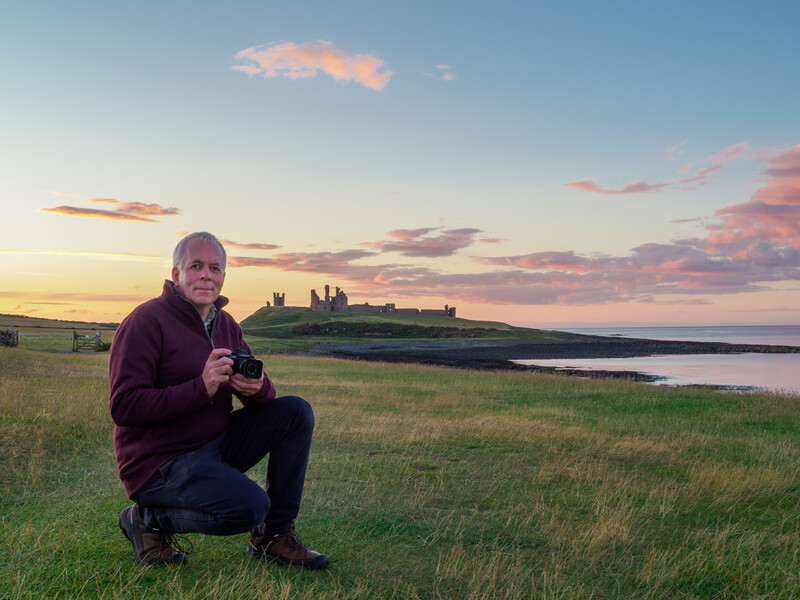 Man crouching in grassy field at sunset with coastal ruins silhouetted across water in background.