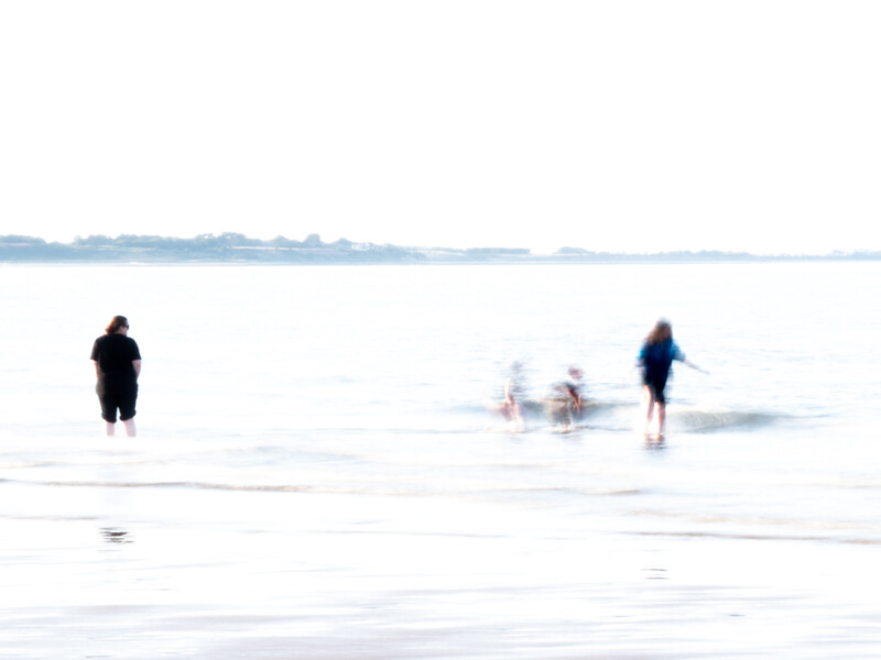 Several people wading in shallow ocean water on an overcast day with a hazy coastal horizon.