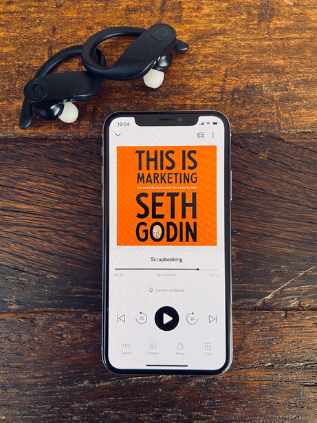 Smartphone displaying audiobook cover for 'This Is Marketing' by Seth Godin, surrounded by wireless earbuds on wooden surface.