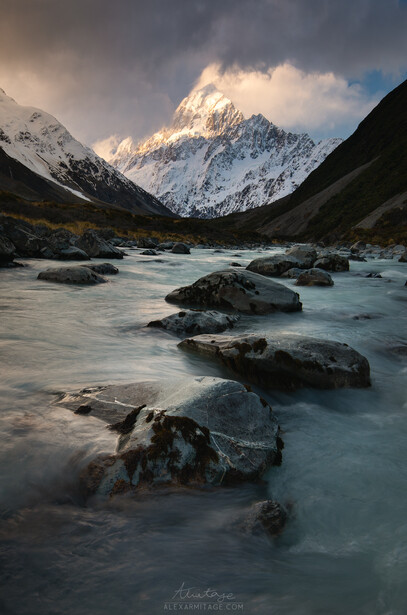 Snow-capped mountain peak rises above a glacial river with scattered boulders in the foreground.