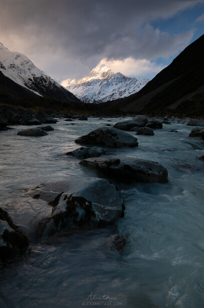 Snow-capped mountain framed by forested valleys with a flowing river dotted with boulders in the foreground.