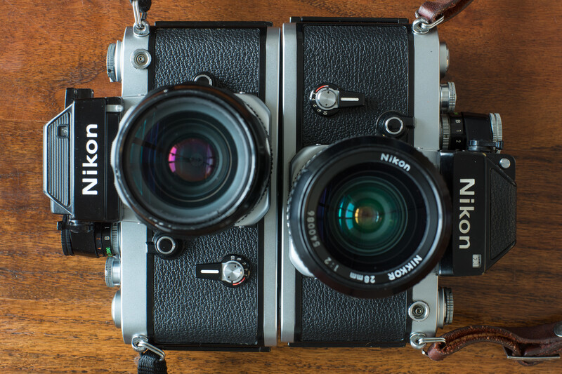 Two Nikon film cameras mounted side-by-side on a wooden surface, each fitted with different lenses.