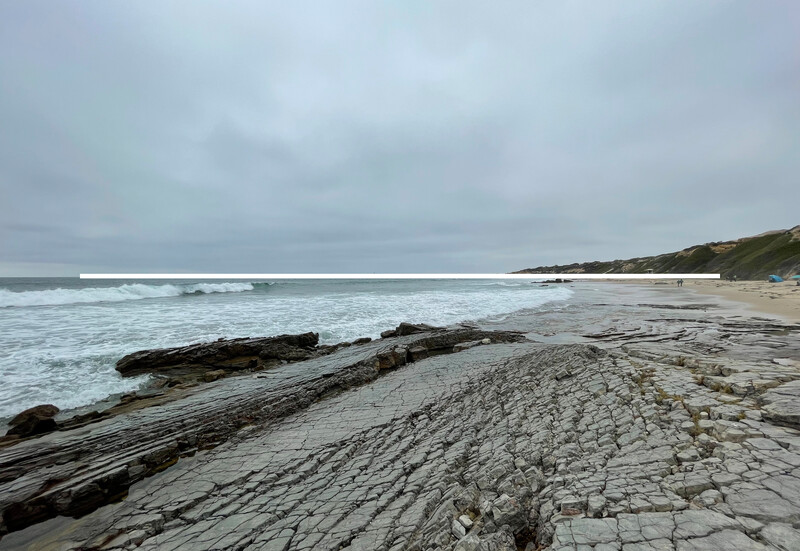 Weathered driftwood log on sandy beach with rocky outcrop and overcast sky.