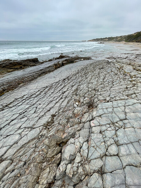 Weathered rock formations with geometric cracks on a beach, waves and sandy shore in background.