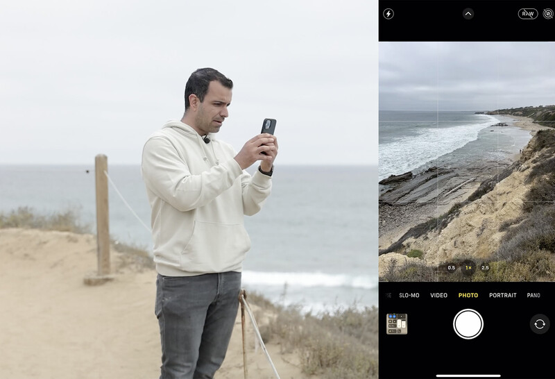 Man holding smartphone to photograph coastal cliffs and beach during overcast conditions.