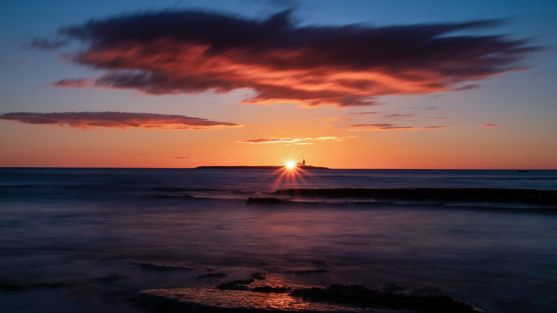 Ocean sunset with dramatic orange and red clouds reflecting on calm water and rocky shore.