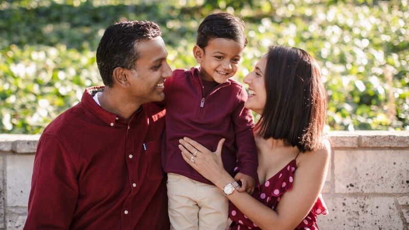 Family of three in burgundy and maroon clothing laughing together outdoors with flowering vines in background.